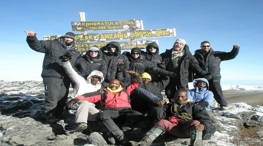 A group of climbers and local guides celebrating at the Uhuru Peak sign (5,895m) on Mount Kilimanjaro after successfully reaching Africa's highest summit during a guided trekking expedition in Tanzania.